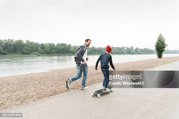 happy father running next to son on skateboard at the riverside - father longboard stock pictures, royalty-free photos & images