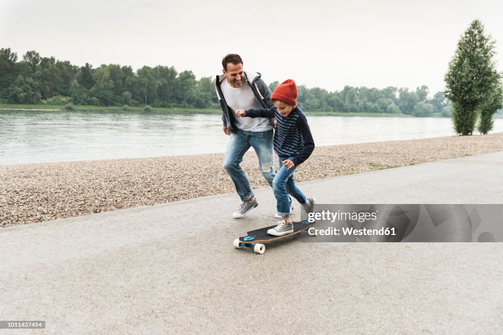 Happy father running next to son on skateboard at the riverside