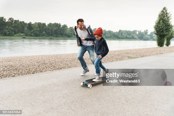 happy father running next to son on skateboard at the riverside - figlio maschio foto e immagini stock