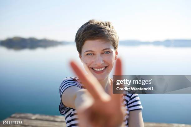 portrait of smiling woman in front of lake showing victory sign - victory sign stock-fotos und bilder