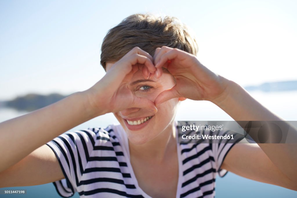 Portrait of smiling woman in front of lake shaping heart with her fingers