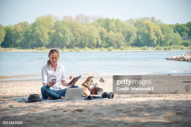 woman sitting on blanket at a river with dog using portable devices - smartphone strand stock-fotos und bilder