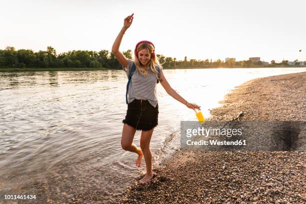 young woman walking barefoot on riverside, earphones and smartphone - wasserrand stock-fotos und bilder