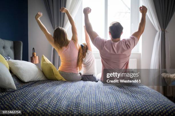 happy family sitting on bed, stretching, rear view - three people bed stock pictures, royalty-free photos & images