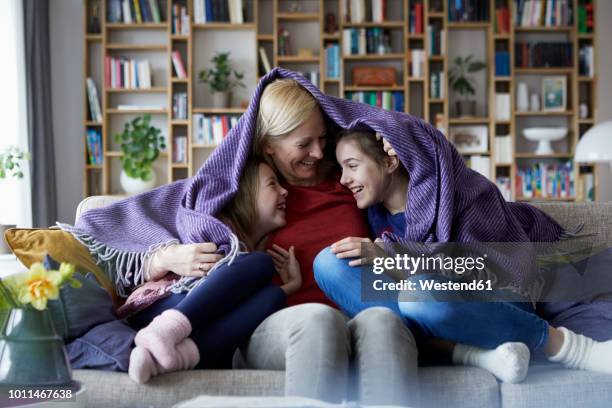 mother and her daughters cuddling and having fun, sitting on couch, covered in blanket - family on sofa stock-fotos und bilder