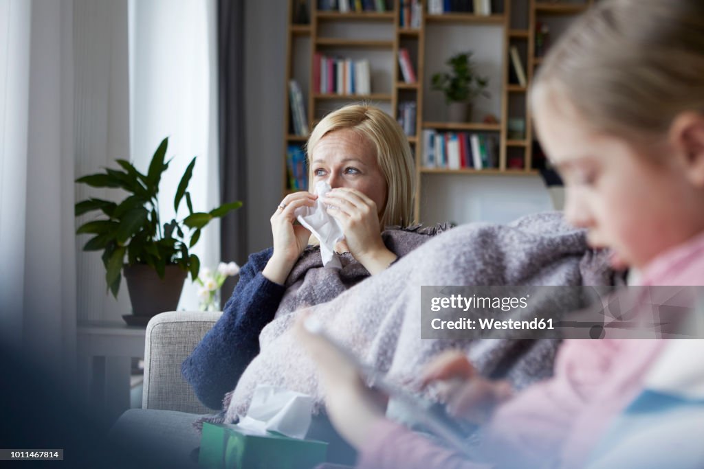 Mother sitting on couch, having a cold, daughter playing in foreground