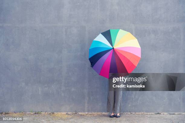 young woman standing with multicolored umbrella in front of a concrete wall - multi coloured umbrella stock pictures, royalty-free photos & images