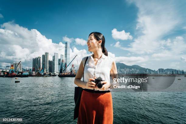 smiling young woman photographing with camera against iconic city skyline and busy commercial dock on a sunny day - asia skyline stock pictures, royalty-free photos & images