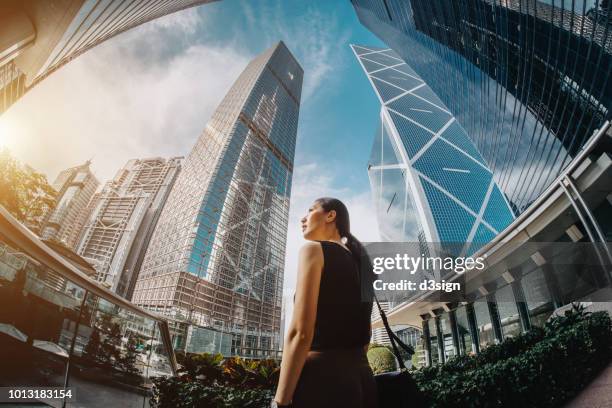 professional asian businesswoman standing against highrise financial towers in central business district and looking up into sky with confidence - em frente de imagens e fotografias de stock