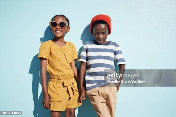 portrait of boy & girl holding hands and looking in camera, on blue backdrop in summer - kids fashion stock-fotos und bilder