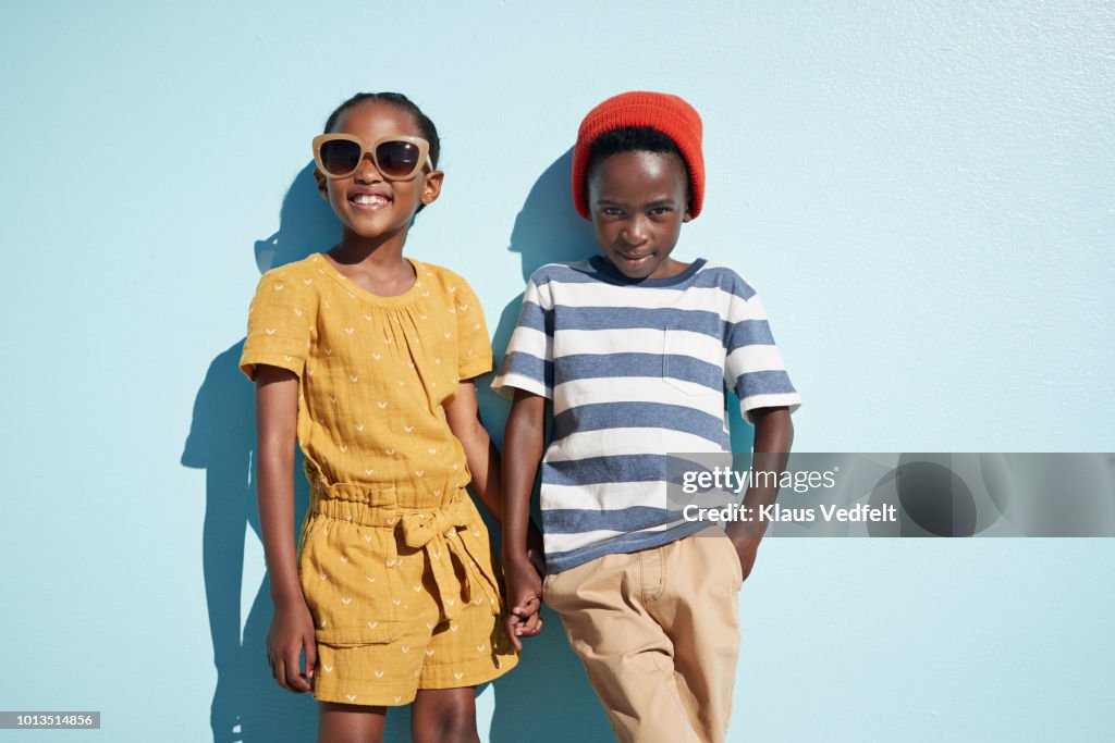 Portrait of boy & girl holding hands and looking in camera, on blue backdrop in summer
