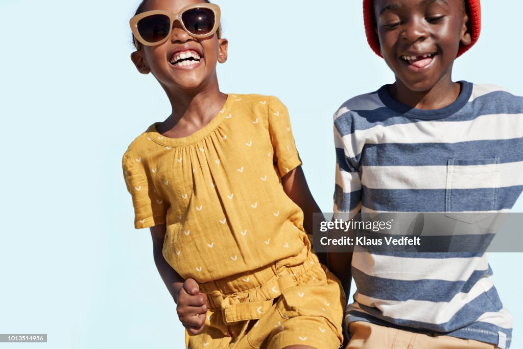 Portrait of boy & girl holding hands and laughing, on blue backdrop in summer