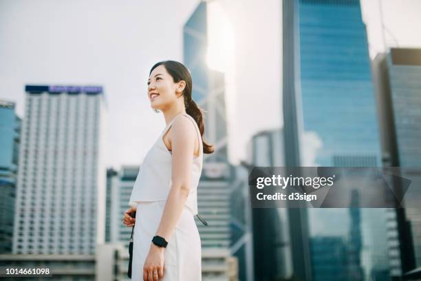 confident young asian businesswoman looking up to sky while standing against urban scene in financial district of hong kong - só mulheres jovens - fotografias e filmes do acervo