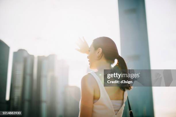beautiful young asian woman shielding eyes from the sun flare while overlooking at city skyline - cutis claro fotografías e imágenes de stock