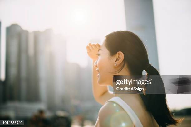 beautiful young asian woman shielding eyes from the sun flare while overlooking at city skyline - blocking sun stock pictures, royalty-free photos & images