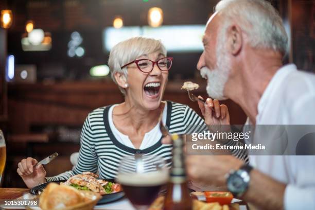 senior woman laughing while feeding her male partner in the restaurant - barbecue social gathering stock pictures, royalty-free photos & images
