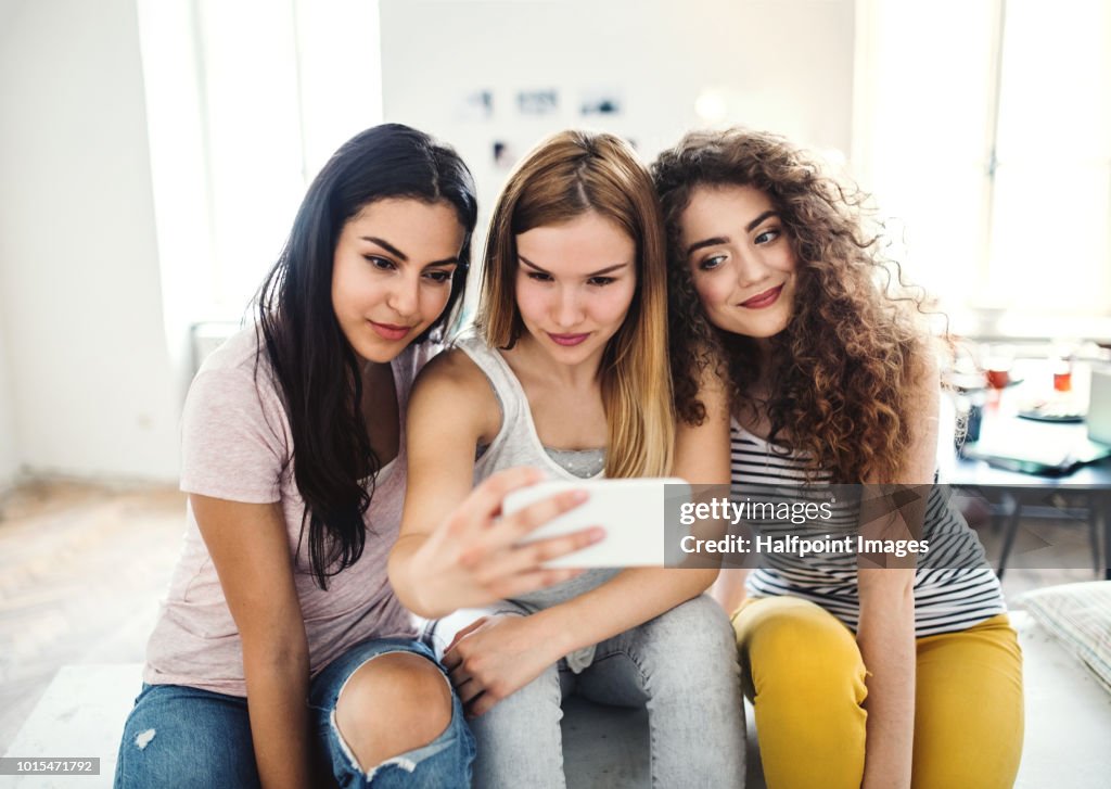 Three female teenager friends with smartphone, sitting at home, taking selfie.