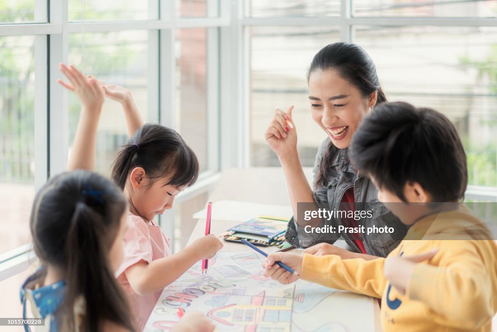 Asian Children boys and girls sitting together around the table in classroom and drawing. With them is their young and beautiful teacher.