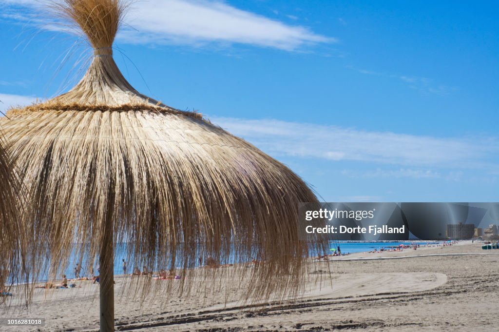 A straw parasol on a sunny beach with a blue sky and some people sunbathing