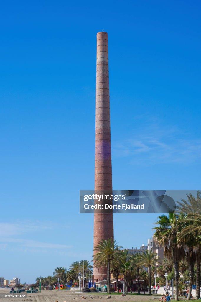 Tall chimney on a beach with some palm trees and sand against a very blue sky