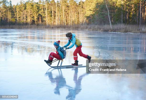 boy pushing his brother on sleigh across frozen lake - family sport winter stock-fotos und bilder