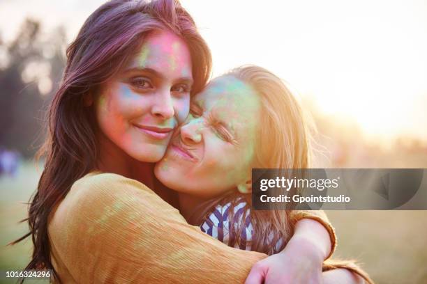 two young women with faces covered in coloured chalk powder hugging at holi festival, portrait - ホーリー祭 ストックフォトと画像