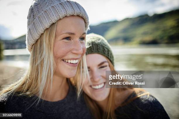 portrait of two smiling female friends wearing knit hats at the riverbank - knit hat stock pictures, royalty-free photos & images