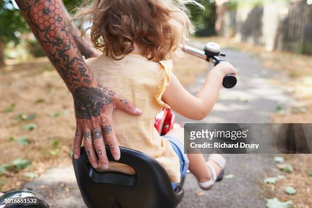 dad helping toddler daughter to ride her tricycle - familie schutz stock-fotos und bilder