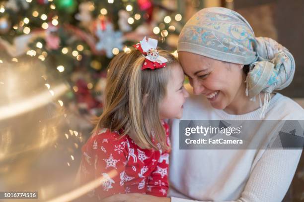 a beautiful mom battling cancer snuggles with her daughter by the christmas tree - christmas treat stock pictures, royalty-free photos & images