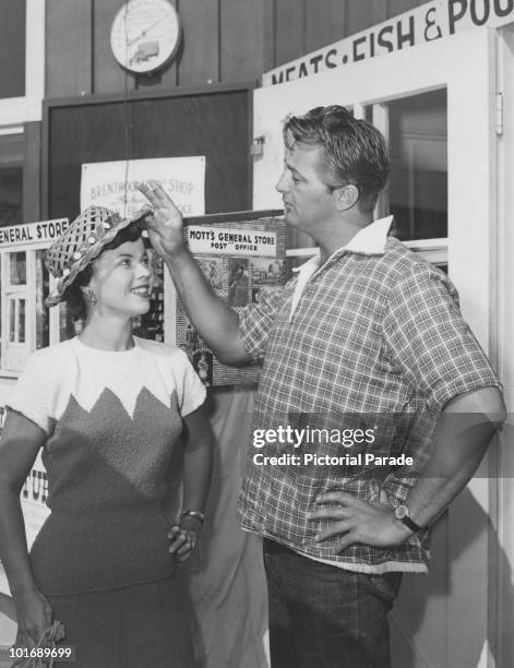 American actress Shirley Temple smiles as fellow actor Robert Mitchum adjusts the brim of her hat while the pair stand in front of miniature 'Mott's...