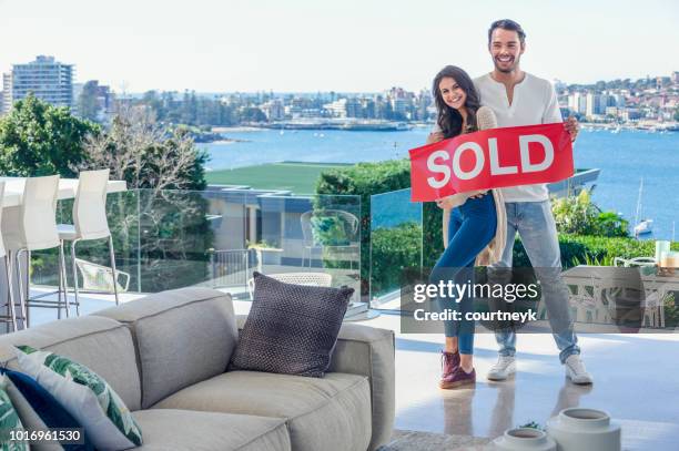 young couple holding a sold sign in a luxury home. - real estate sold stock pictures, royalty-free photos & images