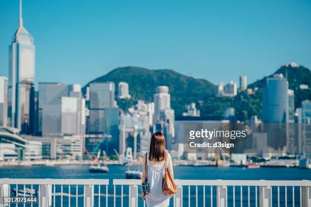 rear view of young woman standing against the promenade of victoria harbour overlooking the cityscape of hong kong, focusing on the foreground - 見渡す ストックフォトと画像