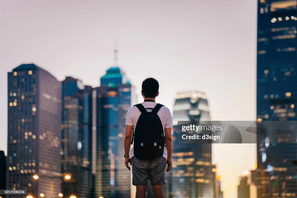 Rear view of man standing in front of illuminated city skyline of Hong Kong at dusk