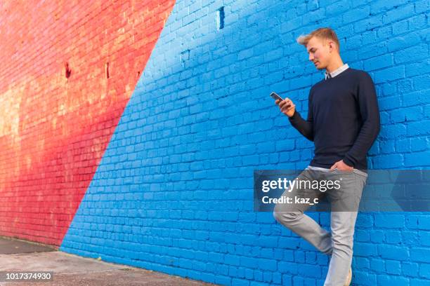 young man leaning against a brick wall while using a smart phone - phone leaning stock pictures, royalty-free photos & images