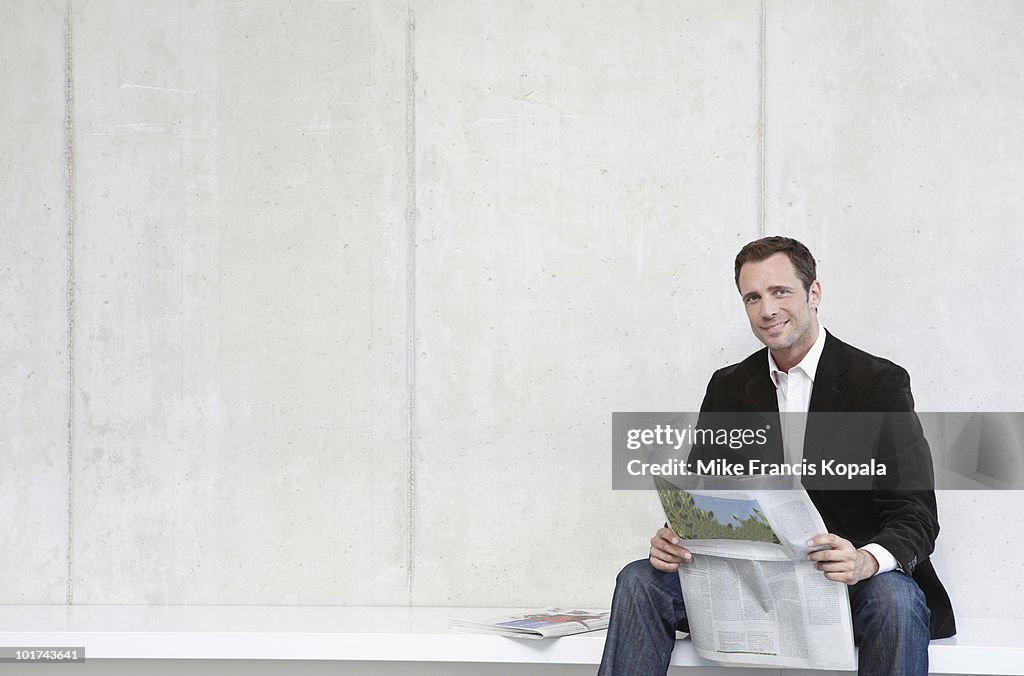 Germany, Cologne, Businessman sitting on bench holding newspaper, smiling, portrait