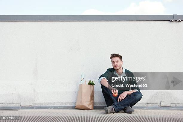 germany, berlin, young man sitting in front of wall, holding an apple, portrait - gambe incrociate foto e immagini stock