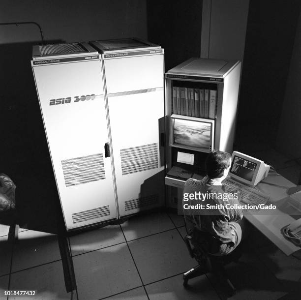 Black and white photograph of Chris Sweeney, from the back, seated at the console of the ESIG-300 Computer, which is used for VMS graphics creation,...
