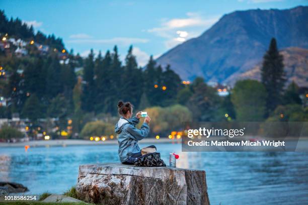 a woman sits on a log lakeside in queenstown on the edge of lake wakatipu and writes in her diary. - queenstown stock pictures, royalty-free photos & images