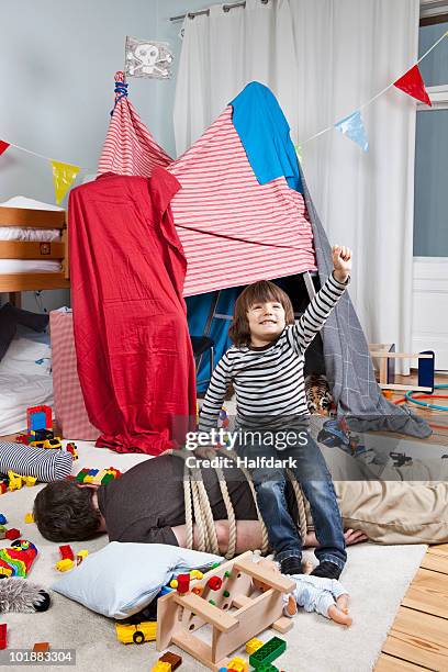 a young boy sitting on a tied up man in a child's playroom - messy playroom stock-fotos und bilder