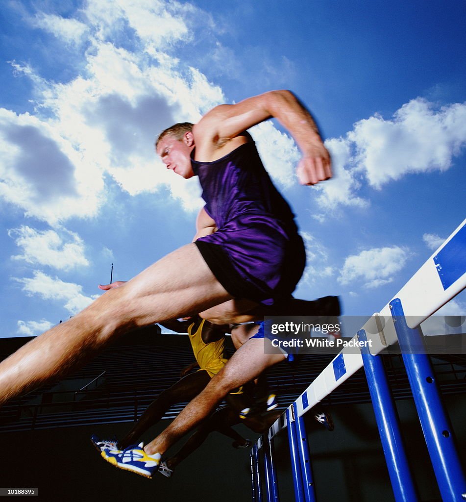 Male athletes leaping hurdles in track and field race, low angle.