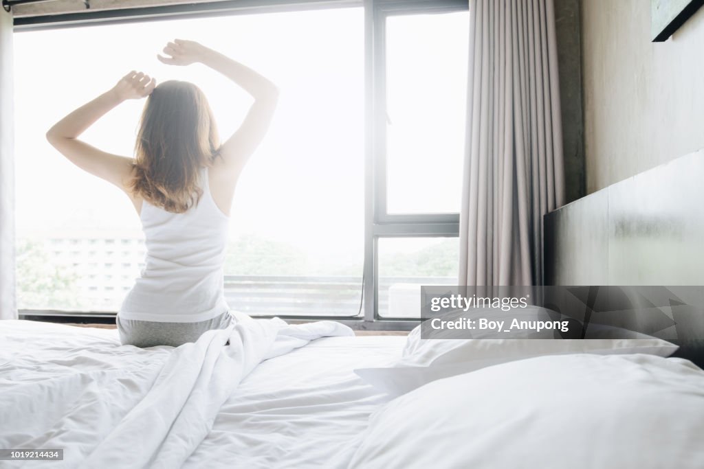 Back view of woman raised hands up and stretching in bed after waking up in the morning, start a new day with happiness.