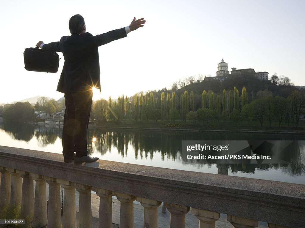 Businessman stands on bridge above river, arms out