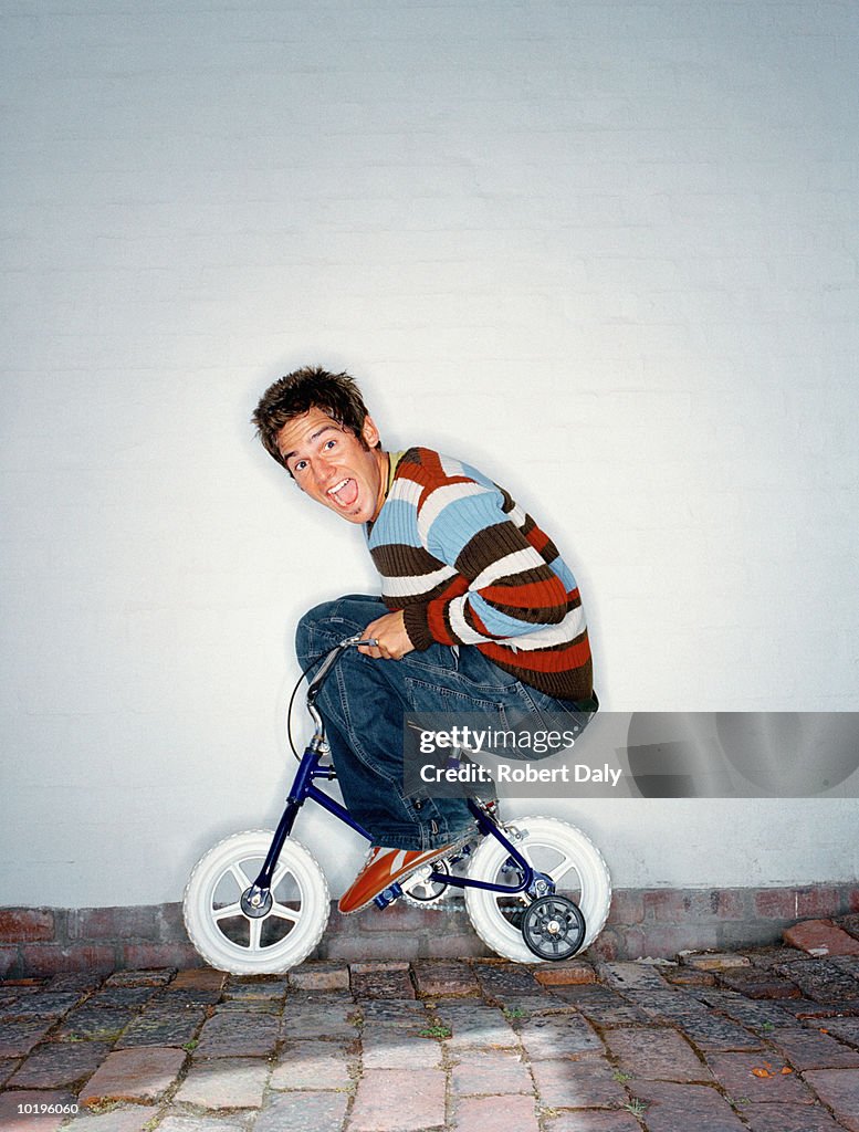 Man riding child's bicycle with stabilisers, mouth open, portrait