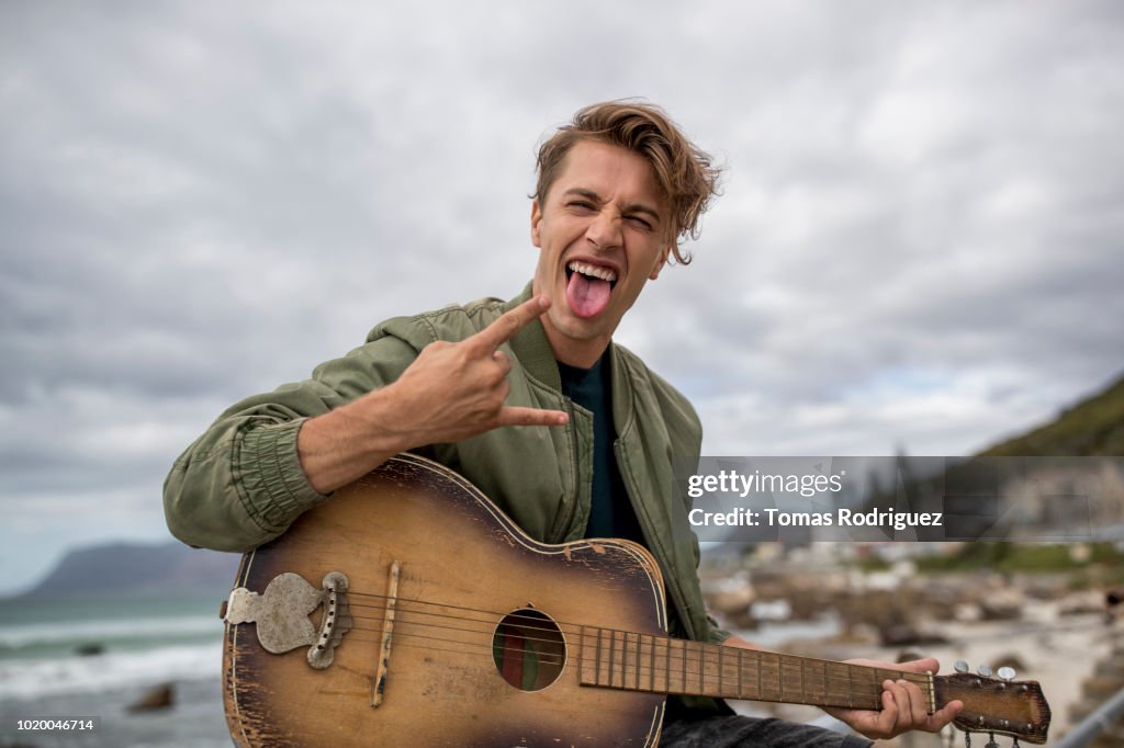 Portrait of carefree young man with guitar at the coast making horn sign