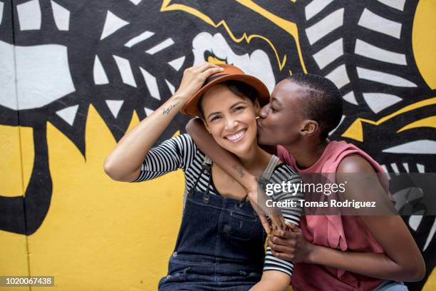 young woman kissing girlfriend in front of graffiti wall - gay couple kissing fotografías e imágenes de stock