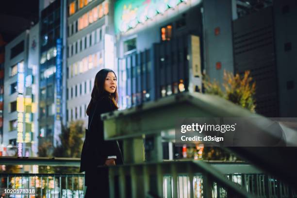 Young woman standing on urban bridge looking over shoulder, against illuminated city buildings in busy Tokyo downtown