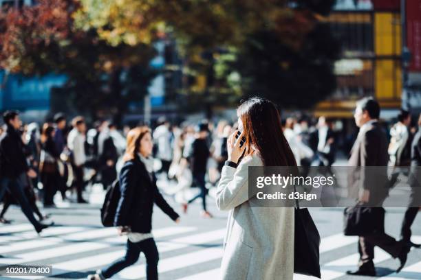 young woman talking on mobile phone while crossing street in downtown district, against busy commuters and city buildings - hora punta temas fotografías e imágenes de stock