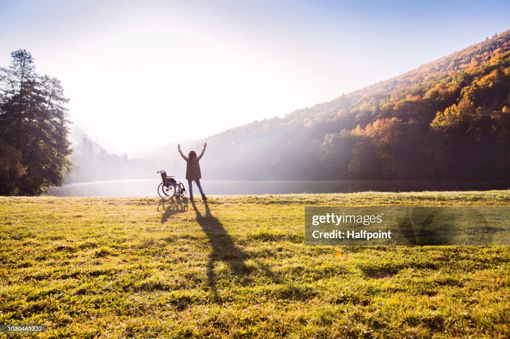 Senior woman standing by the wheelchair in autumn nature.
