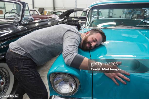 a caucasian male hugging the hood of his old sedan in a classic car repair shop. - voiture de collection photos et images de collection