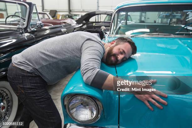 a caucasian male hugging the hood of his old sedan in a classic car repair shop. - hobbies stockfoto's en -beelden